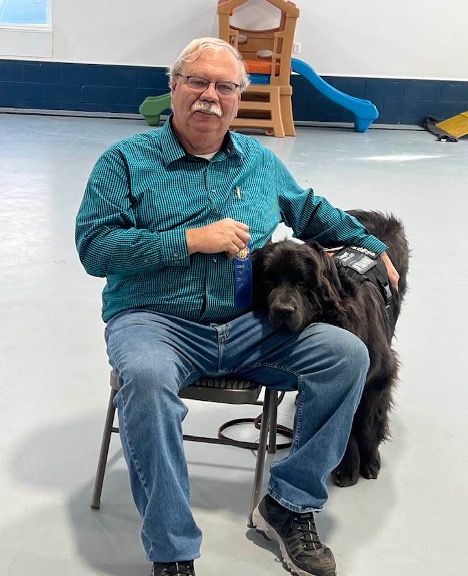 A black Newfoundland with his owner a man with grey hair, a mustache and glasses.